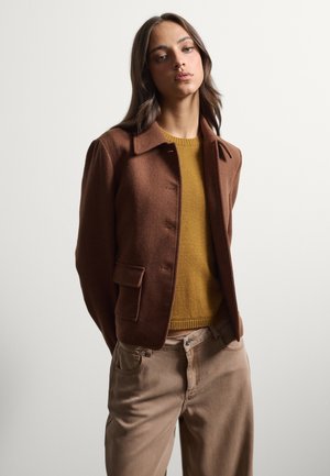 Young woman wearing a brown jacket, mustard yellow sweater, and beige pants, standing with hands behind back against a plain background.