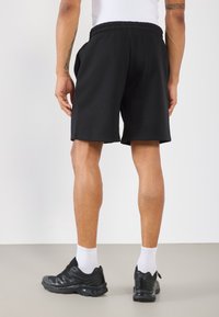 Juodi šortai su elastinga juosta, šoninėmis kišenėmis ir laisvu kirpimu; vilkimi su juodais sportiniais batais ir baltais ankletais.