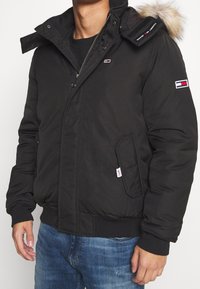 Man som bär svart vinterjacka med pälskantad huva, dragkedja och tryckknappar, två framfickor och blå jeans.