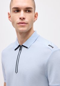 Jeune homme aux cheveux courts portant un polo bleu clair texturé avec une fermeture éclair noire et le logo « BOSS » sur l'épaule, regardant sur le côté.