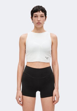Ung kvinde med kort mørkt hår iført en hvid ærmeløs cropped top og sorte atletikshorts med høj talje, stående mod en enkel baggrund.