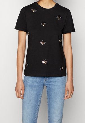 Zwart T-shirt met korte mouwen met geborduurde paillettenbijen, gedragen met lichtblauwe jeans op een persoon die staat tegen een effen achtergrond.