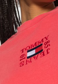 Felpa corallo con scollo a coste, caratterizzata da un ricamo con il testo "TOMMY JEANS" e un logo a bandiera in tonalità rosso, bianco e blu.