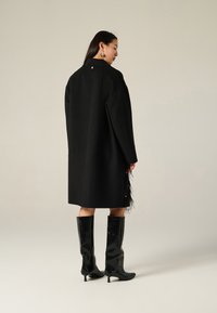 Cappotto di lana nero con vestibilità ampia, dettaglio di frange laterali e spalle strutturate, abbinato a stivali neri lucidi al ginocchio.
