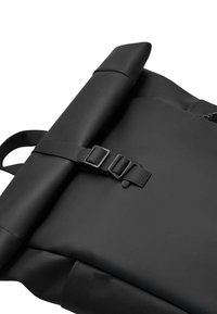 Sac à dos noir à rabat fabriqué à partir d'un matériau lisse et durable avec une fermeture à boucle, présentant un design minimaliste et des bretelles réglables.