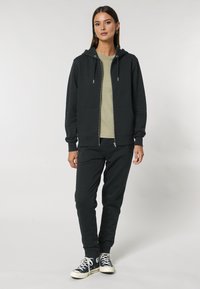 Fekete cipzáras kapucnis pulóver zsinórral, hozzá illeszkedő fekete melegítőnadrággal és világoszöld pólóval. A Converse cipők teszik teljessé a szettet.