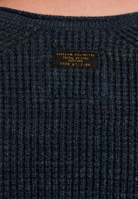 Dzianinowy sweter w ciemnoniebieskim kolorze z teksturowanym wzorem ściągacza. Posiada czarną metkę z złotym napisem "FREEDOM UNLIMITED" oraz szczegółami dotyczącymi pielęgnacji.