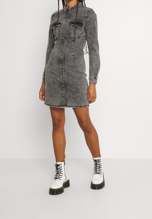 Kobieta ubrana w wyblakłą czarną mini sukienkę z denimu zapinaną na guziki, z kieszeniami na piersi oraz białe sznurowane krótkie buty na grubej podeszwie.