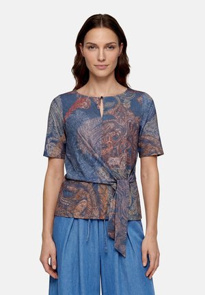 Femme portant un chemisier à manches courtes à motif paisley avec un nœud sur le devant, associé à un pantalon large taille haute bleu clair.