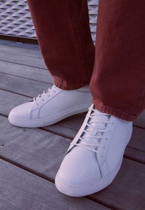 Balti žemi sportbačiai su lygiu odos paviršiumi, su toniniais raišteliais ir guminiais padais, derinami su raudonomis tekstūruotomis audinio kelnėmis.