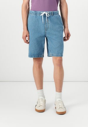 Lichtblauwe denim shorts met een elastische tailleband, witte koordjes en achterzakken, gecombineerd met witte sokken en sneakers.