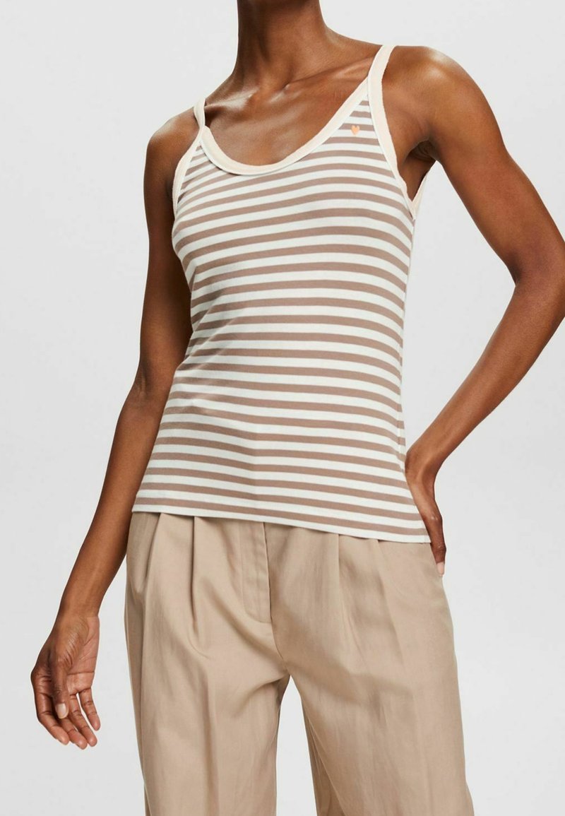 Tunn person klädd i en beige och vit horisontellt randig linne med en liten hjärtdekor och vida beiga byxor mot en enkel bakgrund.