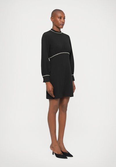 Vestido negro con mangas largas, cuello alto y cintura ajustada, con un ribete decorativo plateado. Combinado con tacones negros.
