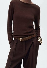 Pull marron côtelé associé à un pantalon marron taille haute, souligné par une ceinture beige et des bracelets dorés au poignet.