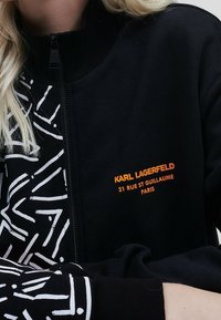 Veste noire zippée avec un motif abstrait blanc audacieux sur une manche et du texte brodé orange portant la mention "KARL LAGERFELD" sur la poitrine.