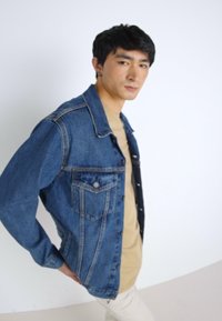 Chaqueta de denim en azul oscuro con botonadura frontal, dos bolsillos y costuras en contraste; llevada sobre una camisa beige, de pie frente a un fondo liso.