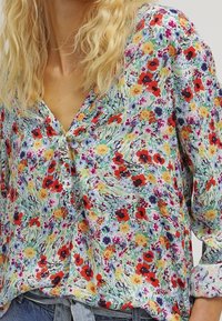 Multicolored floral blouse with a V-neckline, featuring red, yellow, and purple flowers on a light background and button detail at chest.