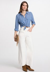Blaues Hemd mit Knöpfen, am Taille gebunden, kombiniert mit weißen weit geschnittenen Hosen. Accessoires umfassen eine beige Fransen-Clutch und braune Stiefeletten.
