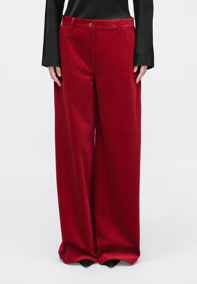 Pantalons larges en velours côtelé rouge avec des côtes verticales prononcées, fermeture à bouton et poches latérales, associés à un haut noir à manches longues.