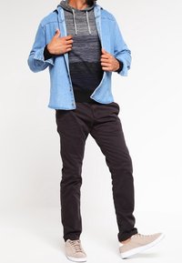 Man die een lichtblauwe denimjack aanpast boven een zwart/grijze hoodie, gekleed in donkere broek en beige sneakers, staande tegen een effen achtergrond.