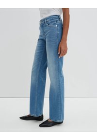 Person klädd i ljusblå raka jeans, vit skjorta och svarta spetsiga ballerinas, stående mot en enfärgad ljusgrå bakgrund.