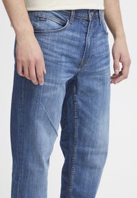 Homme portant un jean en denim bleu clair avec une légère décoloration et une chemise blanche ; mains détendues sur les côtés, visibles de la taille au milieu de la cuisse.