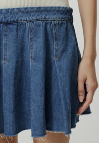 Jeansrok in medium blauw met plooien, een geplooide tailleband en een gerafelde zoom. Eén hand rust op de rok.