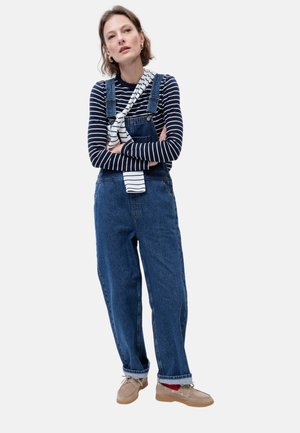 Femme portant une chemise rayée à manches longues et une salopette en jean avec les revers pliés, des mocassins beige et un pull rayé posé sur son épaule.