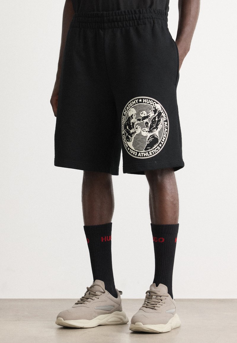 Person kledd i svarte shorts med boksing-grafikk, svarte sokker med rødt "HUGO"-tekst, og beige sneakers som står på et lyst gulv.