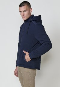 Felpa con zip navy in un tessuto testurizzato, cappuccio con cordino regolabile, tasche laterali, polsini a costine e silhouette aderente.