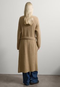 Beige geribde lange cardigan met brede mouwen en een riem in de taille, gecombineerd met loszittende blauwe jeans. Soepele textuur, relaxte pasvorm.