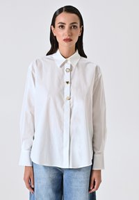 Camicia bianca con colletto classico, dotata di bottoni in metallo dorato e maniche lunghe. Il tessuto appare liscio e leggero.