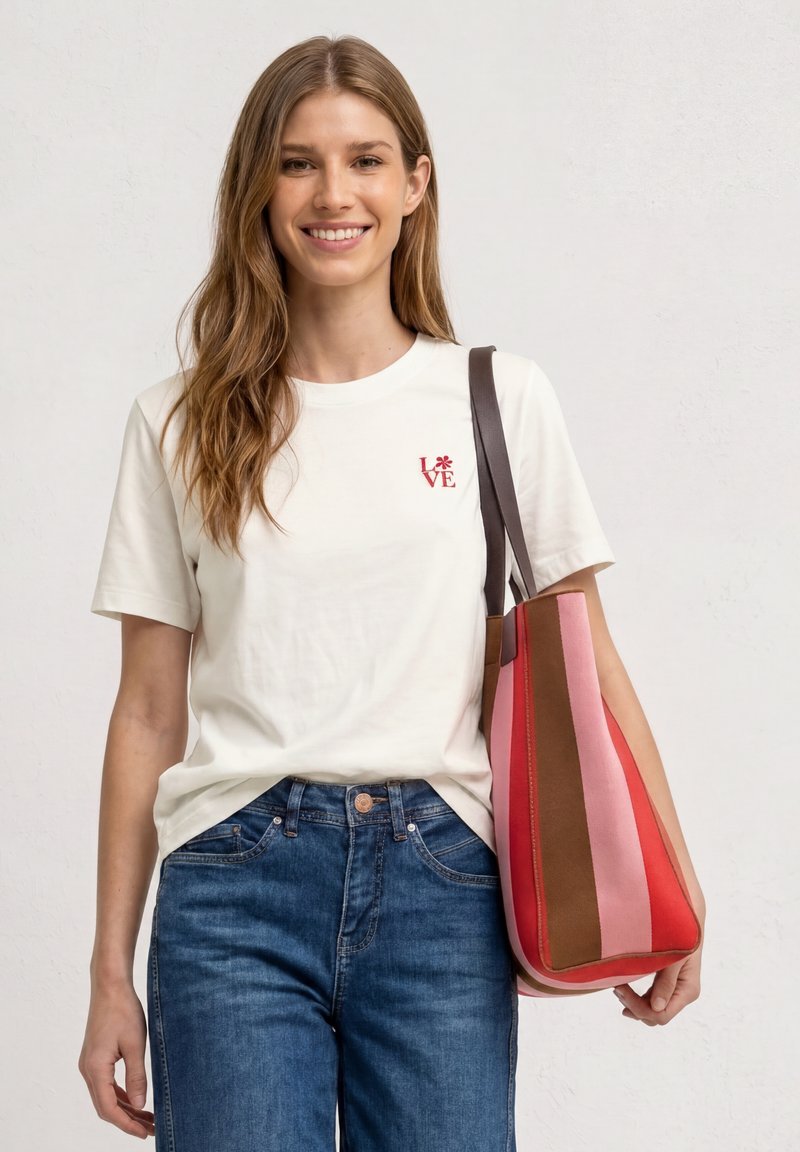 Femme souriante portant un t-shirt blanc "LOVE" et un jean bleu, avec un grand sac à bandoulière rayé rose, rouge et marron sur fond clair.