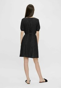 Kobieta z długimi brązowymi włosami, ubrana w czarną sukienkę z krótkim rękawem w białe kropki oraz czarne sandały na płaskiej podeszwie, stoi tyłem.