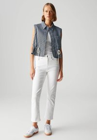 Blauw en wit gestreept mouwloos shirt met koordsluiting in de taille, gecombineerd met witte slim-fit jeans en witte sneakers met beige accenten.