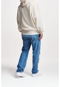 Cremefarbener Kapuzenpullover mit überschnittenen Schultern, kombiniert mit hellblauen, zerrissenen Jeans. Das Model trägt weiße Turnschuhe und steht vor einem neutralen Hintergrund.