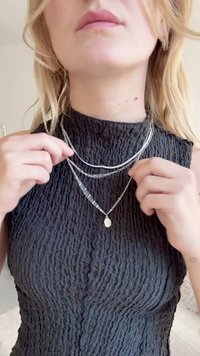 Tri plasti ogrlice iz srebra in metalnih kroglic, z ravnim ovalnim obeskom, razstavljene na teksturirani črni majici.