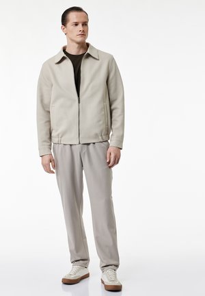 Ung mand står og har en beige lynlåstrøje, mørk skjorte, lysegrå bukser og hvide sneakers med brune såler på.