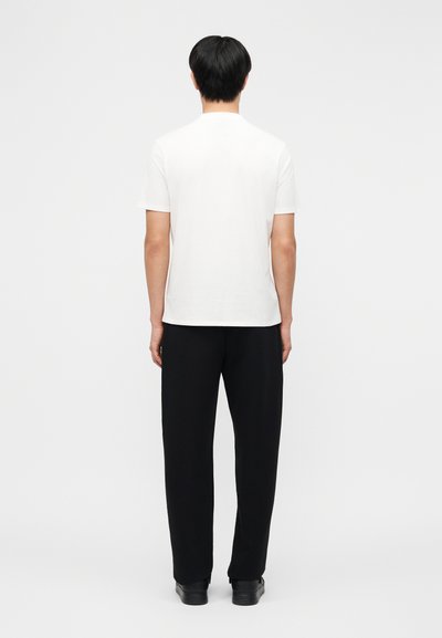 Άνδρας που στέκεται με την πλάτη γυρισμένη, φοράει απλό λευκό κοντομάνικο πουκάμισο, μαύρο παντελόνι και μαύρα παπούτσια μπροστά σε λευκό φόντο.