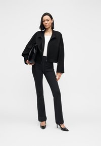 Jachetă neagră croită, cu fermoar și două buzunare frontale, purtată peste un top alb. Pantaloni negri mulați și pantofi stiletto negri cu vârf ascuțit completează ținuta.