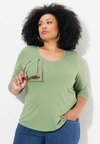 Světle zelená košile s dlouhým rukávem a kulatým výstřihem, měkká textura a volný střih. Model drží sluneční brýle v jedné ruce. Modré džíny.