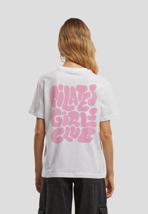 Kvinde med langt bølget hår bærer en hvid t-shirt med stor lyserød tekst "PILATES GIRLS CLUB" på ryggen og sorte cargobukser.