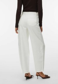 Femme portant un pantalon large blanc taille haute, un haut à manches longues foncé, et des chaussures à talons marron avec des brides à la cheville et des détails en nœud.