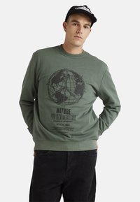 Green sweatshirt featuring a black globe graphic and text "NATURE VS INDUSTRY" on the front. Ribbed cuffs and hem. Cotton blend fabric.