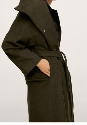 Dark green belted wool coat with wide collar, worn by a person with hand in pocket against plain background.
