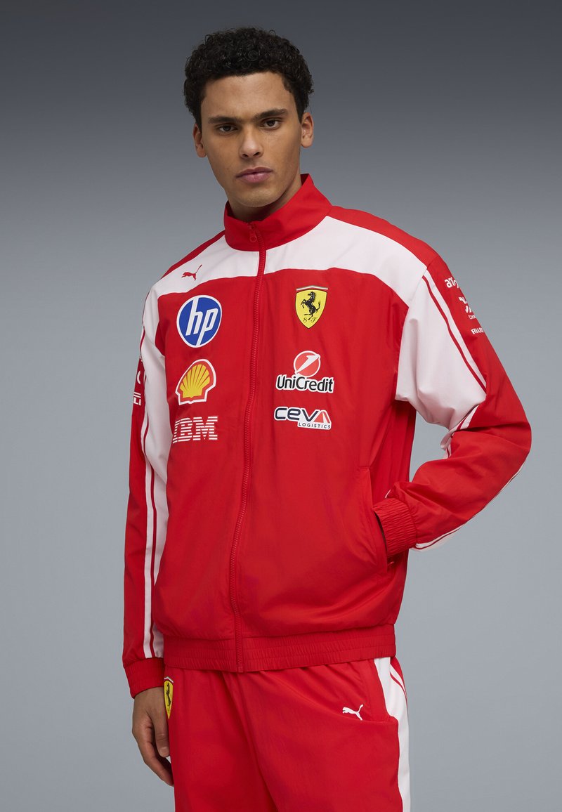 Homme portant une veste et un pantalon de course rouges et blancs avec les logos Ferrari, Shell, HP et Puma, debout devant un fond gris uni.