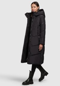 Czarny długi płaszcz puchowy z kapturem, pikowany design, zamek błyskawiczny z przodu i napy. Stylizowany z czarnymi botkami za kostkę i dopasowanymi czarnymi spodniami.