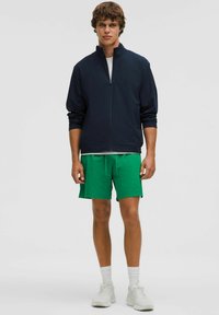 Jūrų laivyno zip-up šalikas, žaliai šortai ir balti kojinės. Batai yra šviesiai pilki. Apranga pasižymi kasdieniu sportiniu dizainu ir kvėpuojančia medžiaga.
