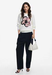 Pulover alb cu un design în formă de floare roz, asociat cu pantaloni negri ajustați și o poșetă crem cu modele decupate. Tocuri negre.