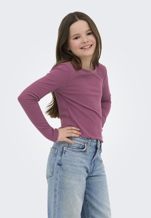 Ung pige med langt brunt hår, smilende, iført en lilla langærmet trøje og lyseblå jeans, står med hænderne på hofterne.
